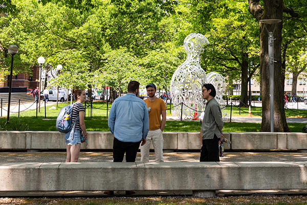 Four people stand facing each other in MIT campus. A large white sculpture and several green trees are in the background