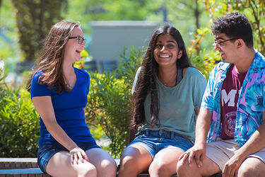 Three smiling MIT students wearing summer clothing and sitting on an outdoor bench with greenery in the background