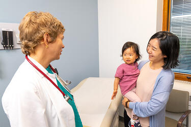 patient holding toddler-age child stand talking with a clinician wearing a lab coat in a medical exam room