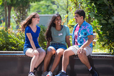 Three smiling MIT students wearing summer clothing sit facing each other on an outdoor bench on MIT campus