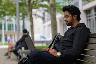 A person sits on an outdoor bench on teh MIT campus while typing on a laptop.