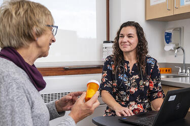 A clinician sits at a table with a laptop across from a patient who holds a prescription bottle