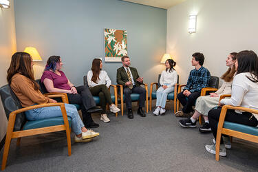 several college students and a clinician sit in chairs arranged in a semi-circle for a group counseling meeting