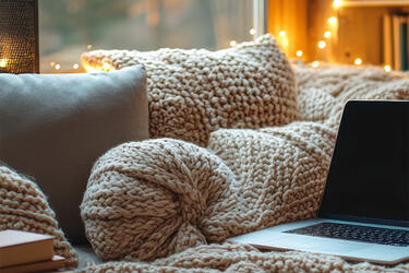 A sofa with several pillows and a blanket with a bookshelf and window in the background and an open laptop on the sofa.