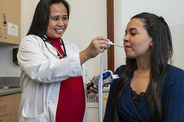 a clinician stand next to a seated patient while taking their temperature with an oral thermometer