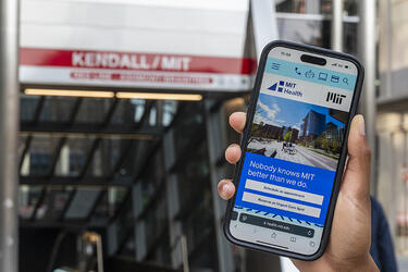 hand holds mobile phone with MIT Health website on the display screen. The MBTA Kendall Square subway station entrance is visible in the background