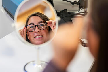 A round table mirror reflects the face of a person trying on eyeglasses. A blurred view of the back of the person’s head and left hand are partially visible in the foreground.