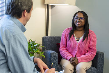 Student seated in a padded armchair faces a partially visible clinician who writes on a clipboard