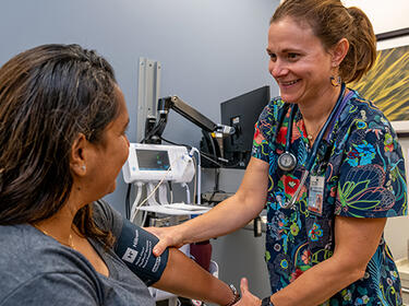 A smiling nurse stands in front of a seated patient while holing a blood pressure cuff around the patient’s right arm