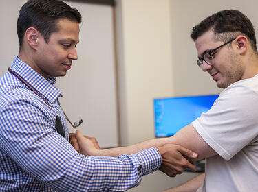 clician stands facing a patient while holding the patient’s forearm and elbow