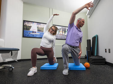 Two people, side by side in a physical therapy room, each stretch their right arm over their head while in a lunging position with their left knee on a support pad, their right knee bent, and their right foot on the floor in front of them