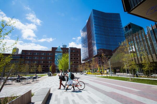 Wide view of MIT Open space plaza on a sunny day, with MIT Health, MIT Building E23, and other campus building visible in the background and two pedestrians in the foreground, one walking a bicycle
