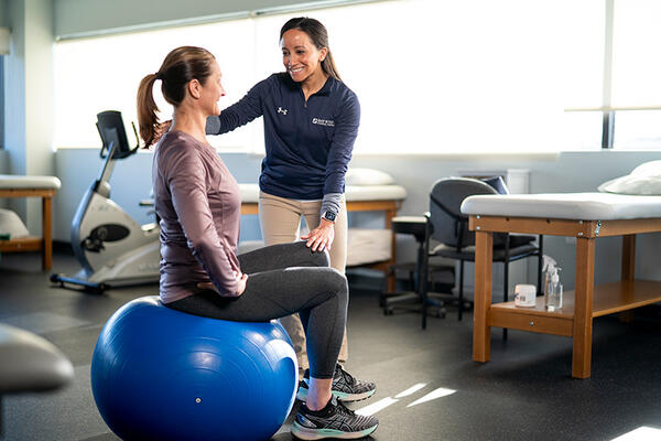 Physical therapist stands next to a patient who sits on a large, inflated therapy ball