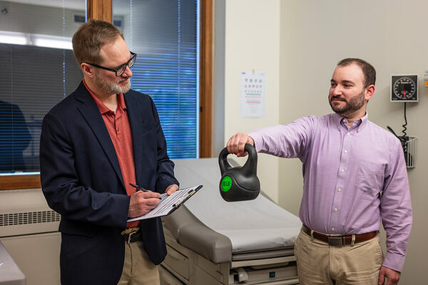 A clinician writes on a clipboard while watching a patient hold up a kettle bell weight with one arm.