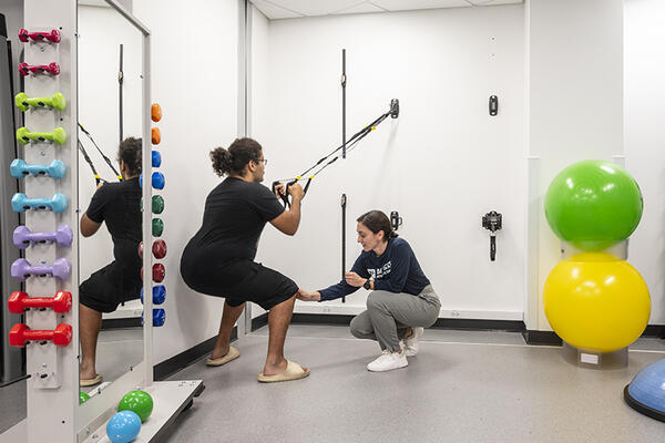 A physical therapist squats next to a patent who is using therapy bands attached to a wall in a physical therapy treatment roomatmnet room 