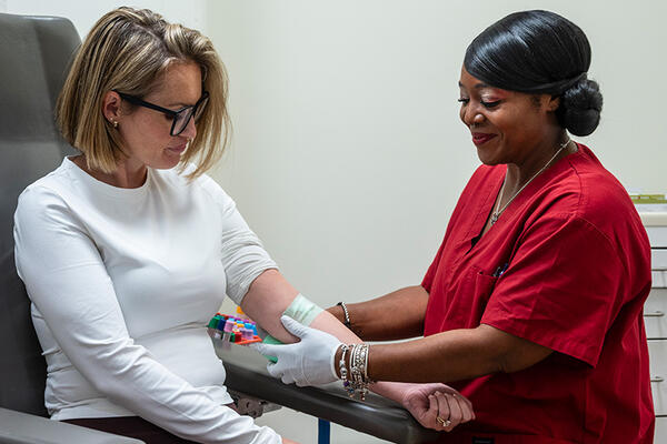 A clinician smiles while pressing a bandage on the inner arm of a patient seated in a phlebotomy chair